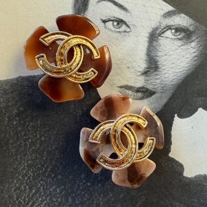 Puede incluir: Dos pendientes con forma de flor con un logotipo de doble C dorado en el centro. Los p&eacute;talos presentan tonos marrones y beige. Los pendientes est&aacute;n colocados sobre una fotograf&iacute;a vintage en blanco y negro.