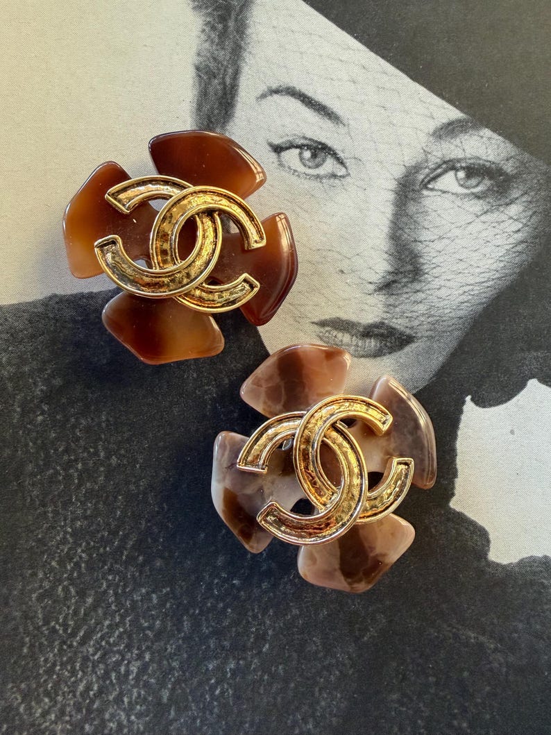 Puede incluir: Dos pendientes con forma de flor con un logotipo de doble C dorado en el centro. Los p&eacute;talos presentan tonos marrones y beige. Los pendientes est&aacute;n colocados sobre una fotograf&iacute;a vintage en blanco y negro.