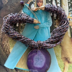 May include: A multi-strand necklace with a large, round, purple agate pendant. The necklace features strands of small, dark beads and gold-colored accents. The pendant is a deep purple with lighter, concentric rings. The necklace is displayed on a vintage background.