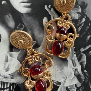 May include: A pair of gold-toned dangle earrings featuring a circular top stud with an intricate embossed spiral pattern. Each earring has a dangling frame with ornate scrollwork accents and two smooth, deep red oval cabochon glass stones.