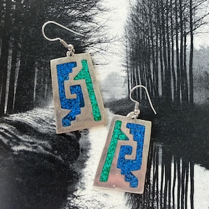 May include: Silver-tone rectangular earrings with inlaid blue and green mosaic designs. The earrings have a hook closure and a geometric pattern. The background is a black and white image of trees.