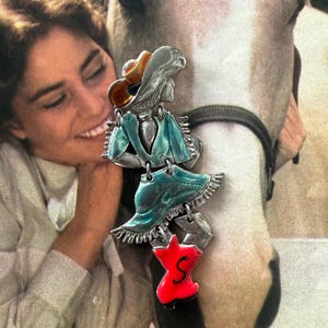 May include: A silver-toned Western-themed pendant with a cowboy hat, turquoise dress, and a red shape with the letter "S". The pendant is hanging in front of a white horse and a person.