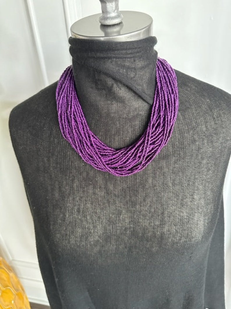 May include: A purple beaded necklace on a black mannequin. The necklace is made of many small beads that are strung together in a long strand.