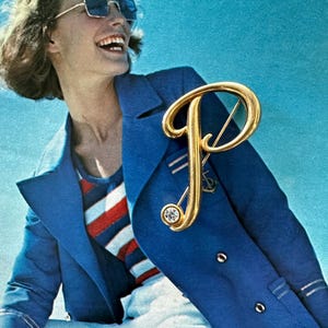 May include: A gold-colored letter "P" brooch with a sparkling crystal accent, pinned to a blue blazer. The blazer is worn over a striped shirt. The image has a vintage aesthetic.
