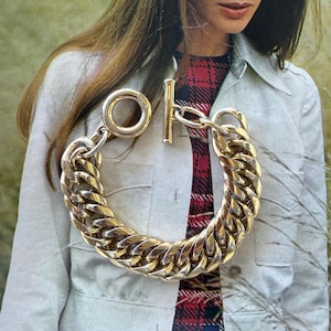 May include: A gold-toned chain bracelet with a large, chunky link design. The bracelet features a T-bar clasp and a large circular loop. The bracelet is shown against a background of a person wearing a light-colored jacket and a plaid dress.
