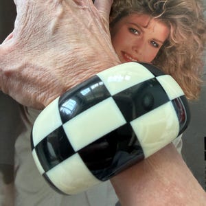 May include: A wide, chunky bangle bracelet featuring a high-gloss finish with a repeating black and off-white checkered pattern. The bracelet has a smooth, rounded rectangular shape that encircles the wrist, showcasing a bold, geometric design.