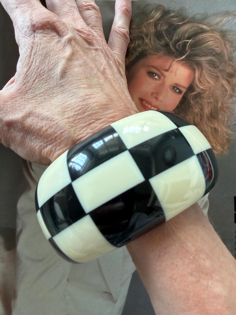 May include: A wide, chunky bangle bracelet featuring a high-gloss finish with a repeating black and off-white checkered pattern. The bracelet has a smooth, rounded rectangular shape that encircles the wrist, showcasing a bold, geometric design.