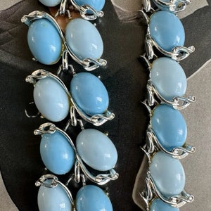 K&ouml;nnte beinhalten: Eine vintage silberfarbene Halskette mit hellblauen ovalen Glasperlen. Die Perlen sind in einem silberfarbenen Metallrahmen mit einem Blattdesign eingefasst.