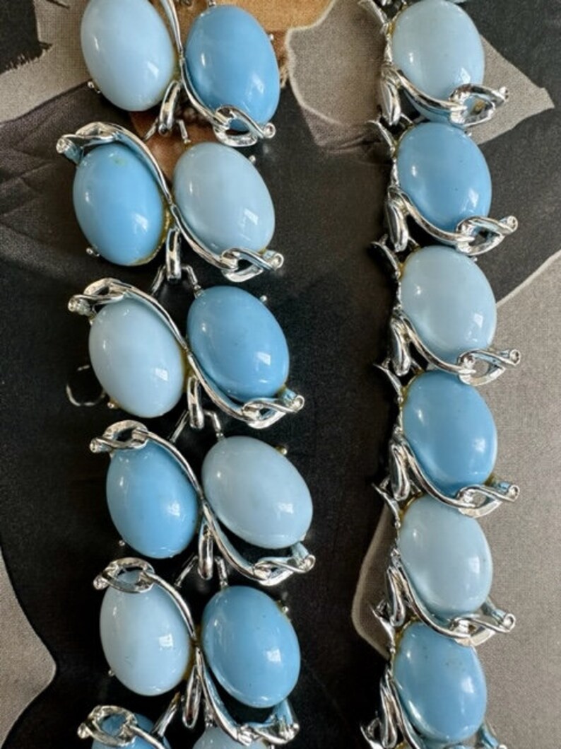 K&ouml;nnte beinhalten: Eine vintage silberfarbene Halskette mit hellblauen ovalen Glasperlen. Die Perlen sind in einem silberfarbenen Metallrahmen mit einem Blattdesign eingefasst.