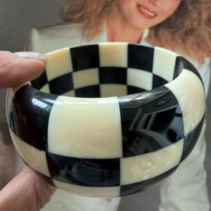 May include: A wide bangle bracelet featuring a high-contrast checkerboard pattern of alternating black and off-white squares. The bracelet has a smooth, glossy finish and a thick, rounded profile that encircles the wrist with a bold geometric design.