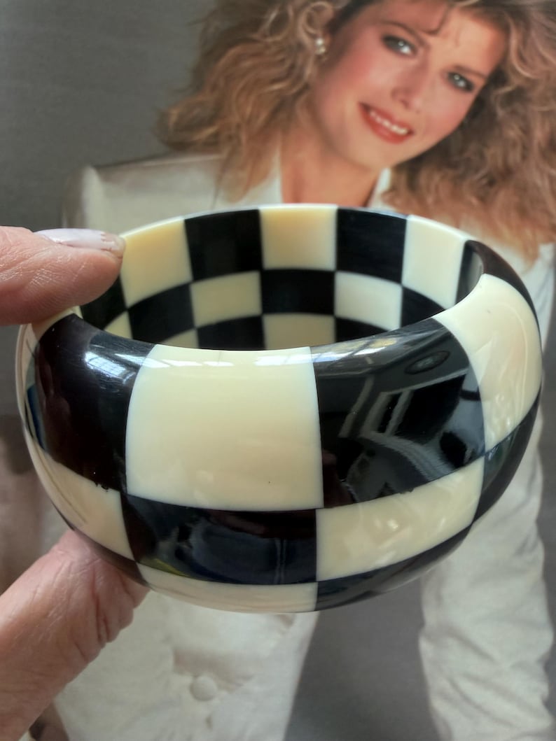 May include: A wide bangle bracelet featuring a high-contrast checkerboard pattern of alternating black and off-white squares. The bracelet has a smooth, glossy finish and a thick, rounded profile that encircles the wrist with a bold geometric design.