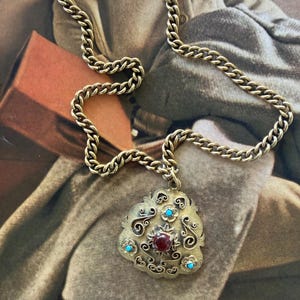 May include: A gold-toned necklace with a decorative pendant. The pendant features a central red stone, surrounded by turquoise-colored stones and ornate scrollwork. The chain is a thick, linked design.
