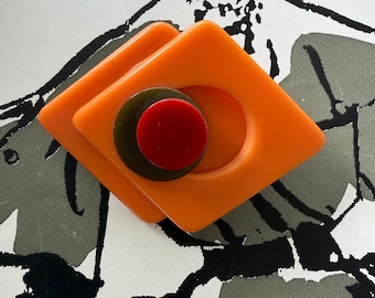 Könnte beinhalten: Eine Vintage-Brosche aus orangefarbenem Bakelit mit geometrischem Design. Die Brosche hat zwei übereinanderliegende rautenförmige Teile mit einem kreisförmigen Ausschnitt und einen dunkelgrünen und roten Kreis in der Mitte. Die Brosche ist vor einem schwarz-weißen Hintergrund.
