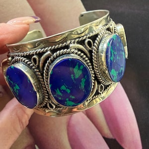 May include: Silver-toned cuff bracelet featuring three oval, deep blue stones with green marbling. The stones are set in ornate, rope-like frames. The bracelet has an open design and is likely made of metal.