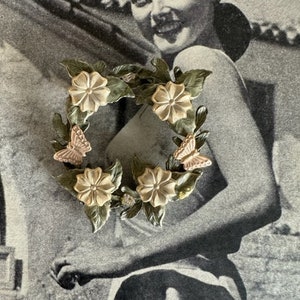 May include: A vintage-style brooch featuring a wreath design with cream-colored flowers, green leaves, and two pink butterflies. The brooch is set against a black and white background, creating a classic and elegant aesthetic.