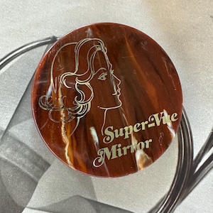 May include: A round, vintage compact mirror with a brown and orange marbled design. The mirror features a profile of a woman's face and the words "Super-Vite Mirror" in gold lettering.