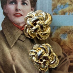 May include: A pair of gold-toned clip-on earrings. Each earring features a woven knot design, reflecting light. The earrings are set against a vintage background with a woman in a brown coat.