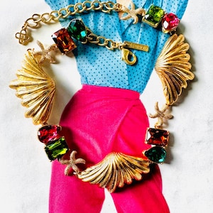 May include: A gold-toned statement necklace featuring a seashell design with colorful gemstones and starfish accents. The necklace has a chain and clasp closure. The gemstones are red, green, and orange. The necklace is displayed on a background of a blue shirt and pink pants.