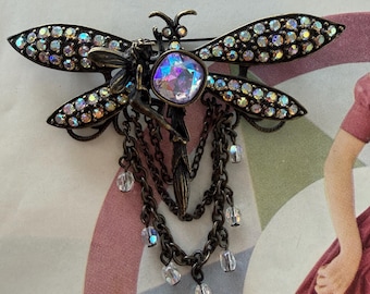 May include: A bronze-toned dragonfly brooch with iridescent rhinestones on the wings and a large, multi-colored crystal at the center. The brooch features a small fairy figure and dangling chains with clear beads.
