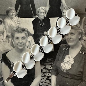 May include: A white vintage bracelet with oval-shaped links and a silver clasp. The bracelet is made of plastic and metal. The bracelet is shown in front of a black and white photo of a group of women.