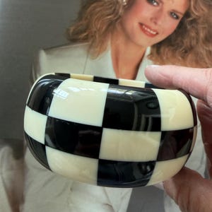 May include: A wide, thick bangle bracelet featuring a high-gloss, alternating black and off-white checkered pattern. The bracelet has a smooth, rounded exterior surface and a substantial, chunky profile with a consistent grid design throughout.