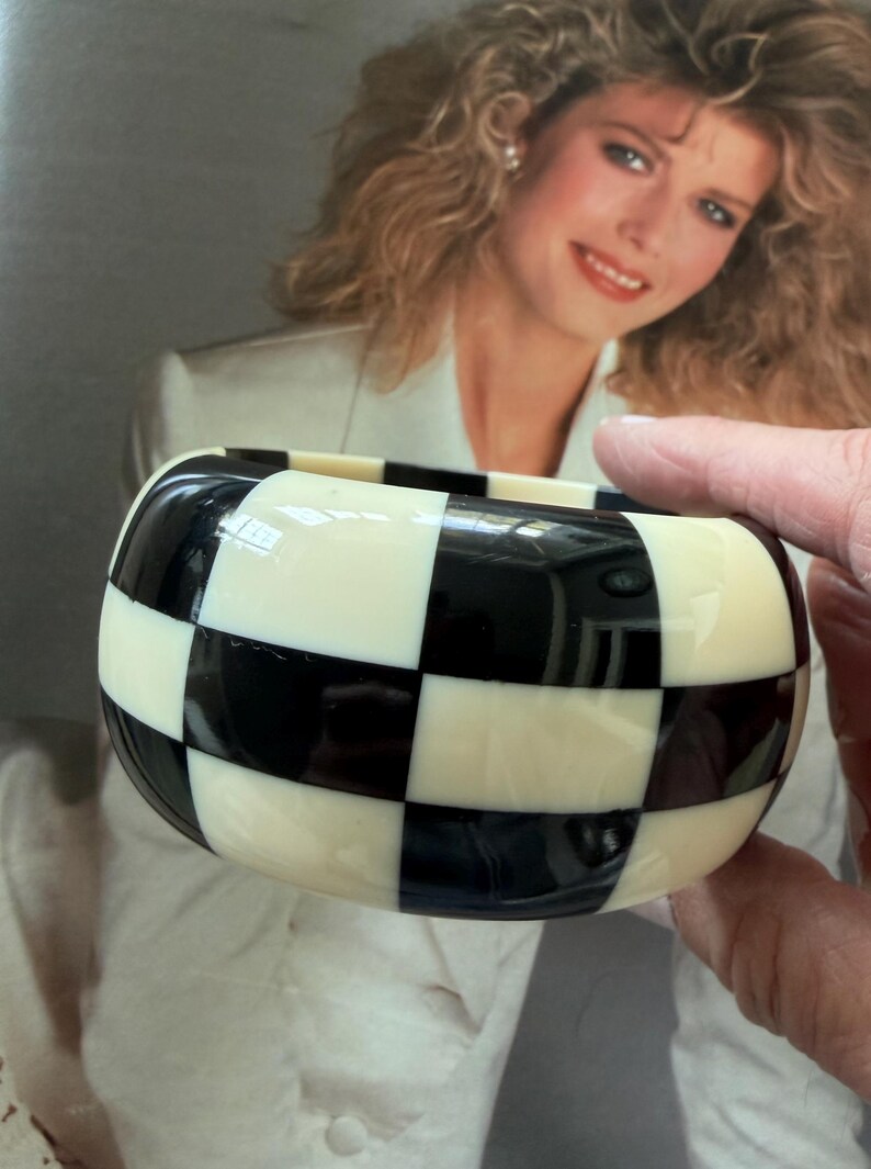 May include: A wide, thick bangle bracelet featuring a high-gloss, alternating black and off-white checkered pattern. The bracelet has a smooth, rounded exterior surface and a substantial, chunky profile with a consistent grid design throughout.