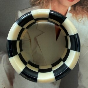 May include: A thick, circular bangle bracelet featuring a high-contrast checkerboard pattern of alternating black and off-white squares. The rigid, smooth-surfaced accessory has a wide band design that forms a complete ring with a glossy finish.