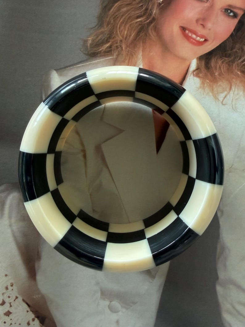 May include: A thick, circular bangle bracelet featuring a high-contrast checkerboard pattern of alternating black and off-white squares. The rigid, smooth-surfaced accessory has a wide band design that forms a complete ring with a glossy finish.