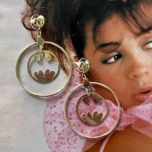 May include: A pair of gold-toned drop earrings featuring a large circular hoop frame. Inside each hoop hangs a smaller circular charm with a cut-out design resembling a stylized bat silhouette. The earrings have a post-back closure mechanism.