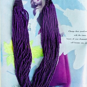 May include: A long purple beaded necklace with a gold button clasp. The necklace is made of many strands of small beads.