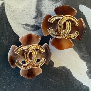 Puede incluir: Dos pendientes marrones y dorados con forma de flor. Cada pendiente presenta un logotipo doble C dorado en el centro. Los p&eacute;talos son una mezcla de marr&oacute;n claro y oscuro. Sobre un fondo en blanco y negro.