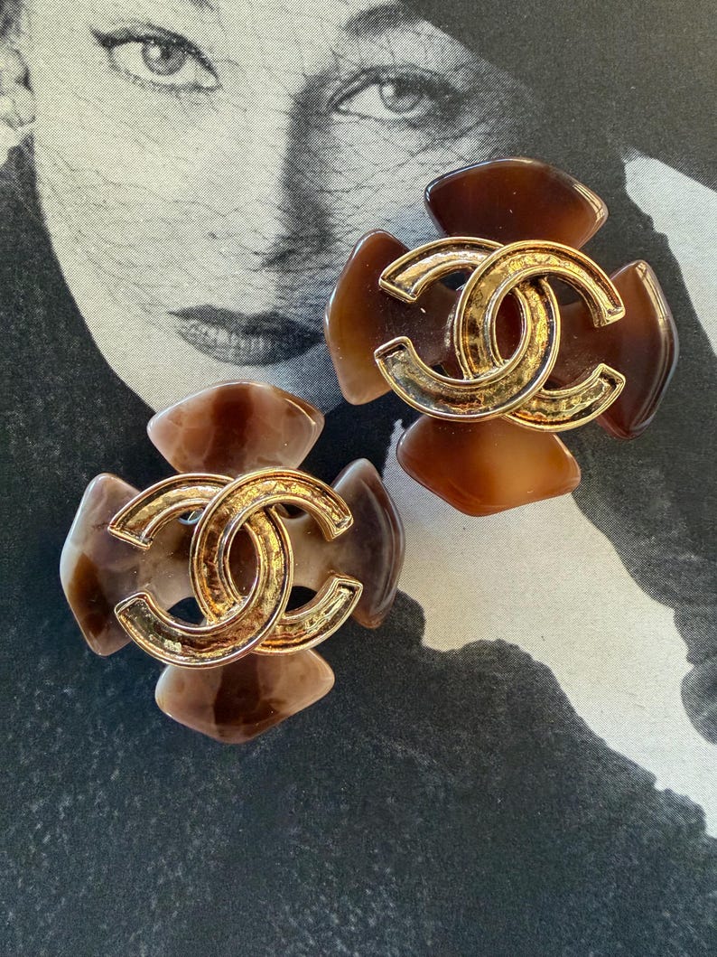 Puede incluir: Dos pendientes marrones y dorados con forma de flor. Cada pendiente presenta un logotipo doble C dorado en el centro. Los p&eacute;talos son una mezcla de marr&oacute;n claro y oscuro. Sobre un fondo en blanco y negro.