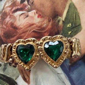 May include: A vintage gold-toned bracelet featuring two heart-shaped emerald green gemstones. The hearts are framed with ornate gold detailing. The bracelet has an expandable design and is set against a vintage-style background.