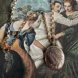 May include: A gold-toned necklace with a detailed oval locket. The locket features an ornate, engraved floral design. The chain is a classic link style. The background is a vintage painting with figures in period clothing.