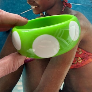 May include: A bright green bangle bracelet with large white polka dots. The bracelet is round and appears to be made of a glossy material. The bracelet is being held up by a person.