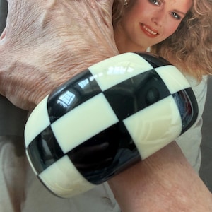May include: A wide, chunky bangle bracelet featuring a high-gloss finish with a repeating black and white checkerboard pattern. The bracelet has a smooth, rounded surface and a thick, bold design that wraps around the wrist with a uniform grid.