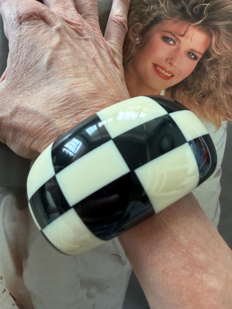 May include: A wide, chunky bangle bracelet featuring a high-gloss finish with a repeating black and white checkerboard pattern. The bracelet has a smooth, rounded surface and a thick, bold design that wraps around the wrist with a uniform grid.