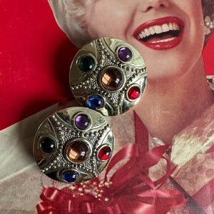 May include: A pair of silver tone earrings with a starburst design and multi-colored gemstones. The earrings have a round shape and are decorated with a beaded border.