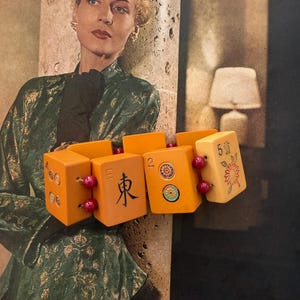 May include: A vintage bracelet made of orange Mahjong tiles, connected by small red beads. The tiles feature Chinese characters and symbols. The background includes a vintage photograph of a woman in a fur hat and green coat.