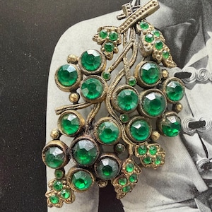 May include: Vintage brooch in the shape of a grape cluster. The brooch features a gold-toned metal frame with a detailed vine design. The grapes are represented by large, faceted emerald green glass stones, adding a touch of sparkle and elegance.