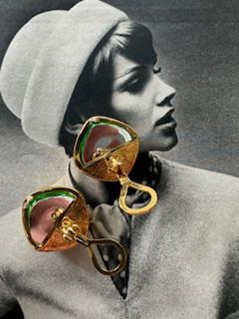 May include: A pair of gold-toned, square earrings with a unique design. Each earring features a central pink element, surrounded by green and gold accents. The earrings have a distinctive handle-like shape, adding to their vintage appeal.