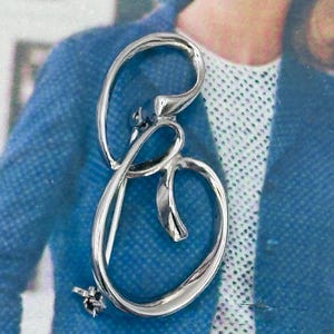 May include: A silver-toned brooch in the shape of the letter "B". The brooch has a flowing, cursive design with a small floral accent at the bottom. The background is a blue and white patterned fabric.