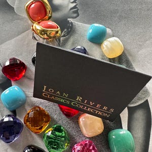 May include: A gold-toned clip-on earring with a coral-colored stone, displayed with a collection of colorful, faceted gemstones. The black box reads "JOAN RIVERS CLASSICS COLLECTION". The gemstones are various colors including red, blue, green, and pink.