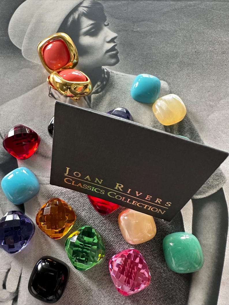 May include: A gold-toned clip-on earring with a coral-colored stone, displayed with a collection of colorful, faceted gemstones. The black box reads "JOAN RIVERS CLASSICS COLLECTION". The gemstones are various colors including red, blue, green, and pink.