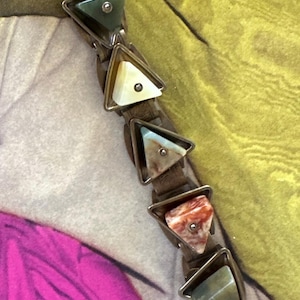 May include: A unique bracelet featuring triangular stones in shades of red, green, and white, set in silver-toned metal. The stones are connected by a brown leather band, creating a striking and artistic accessory.