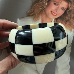 May include: A wide bangle bracelet featuring a high-gloss finish with a repeating black and off-white checkered pattern. The circular accessory has a smooth, rounded exterior surface and a thick, chunky profile that creates a bold visual contrast.