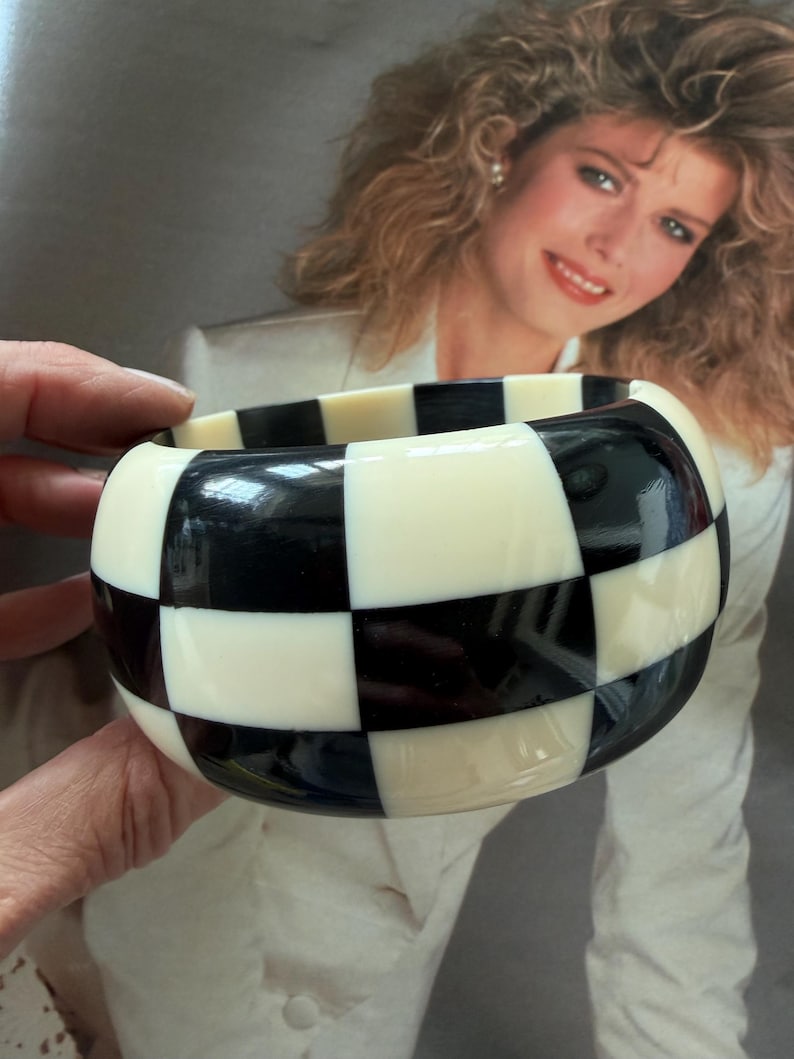 May include: A wide bangle bracelet featuring a high-gloss finish with a repeating black and off-white checkered pattern. The circular accessory has a smooth, rounded exterior surface and a thick, chunky profile that creates a bold visual contrast.