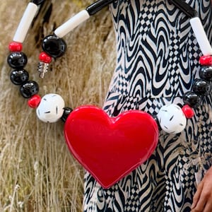 May include: A bold necklace featuring a large red heart pendant. The necklace has black, white, and red beads, with a black and white clasp. The necklace is a statement piece, perfect for adding a pop of color.