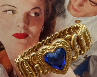 Può includere: Bracciale dorato con una gemma blu a forma di cuore. Il cuore è incorniciato da una cornice dorata ornata. Il bracciale ha un design flessibile con maglie decorative. Lo sfondo presenta un'illustrazione in stile vintage.