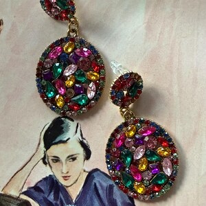 May include: A pair of gold-toned earrings with a large, round, colorful gemstone design. The earrings feature a smaller, matching gemstone cluster at the top. The gemstones are in various shapes and colors, including red, green, pink, and yellow.